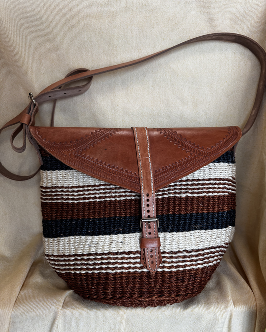 A woven shoulder bag with a brown leather flap and strap, featuring horizontal stripes in brown, cream, and black, resting on a beige fabric background.