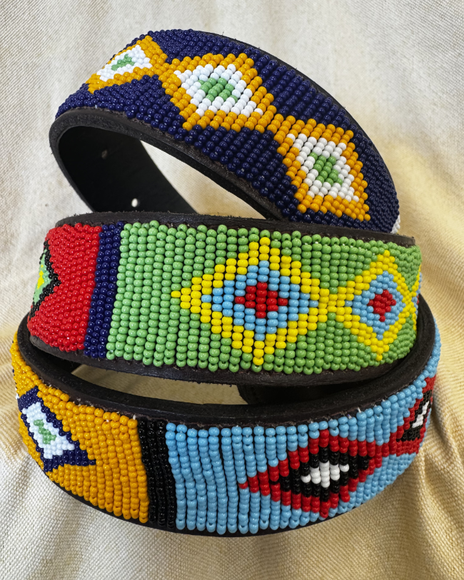Three handmade beaded waist belts with intricate geometric patterns in yellow, green, blue, red, and white, displayed on a beige fabric background.