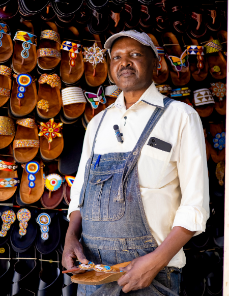 A man wearing a white shirt and blue denim overalls stands in front of a wall displaying many colorful handmade sandals, holding a small tray with a pair of sandals. A smartphone is tucked into his pocket, and a small lapel microphone is clipped to his shirt.